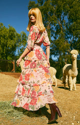 A woman wearing a pink floral tiered maxi dress and dark brown heeled boots stands outdoors in a sunny farm setting with sheep and trees in the background.